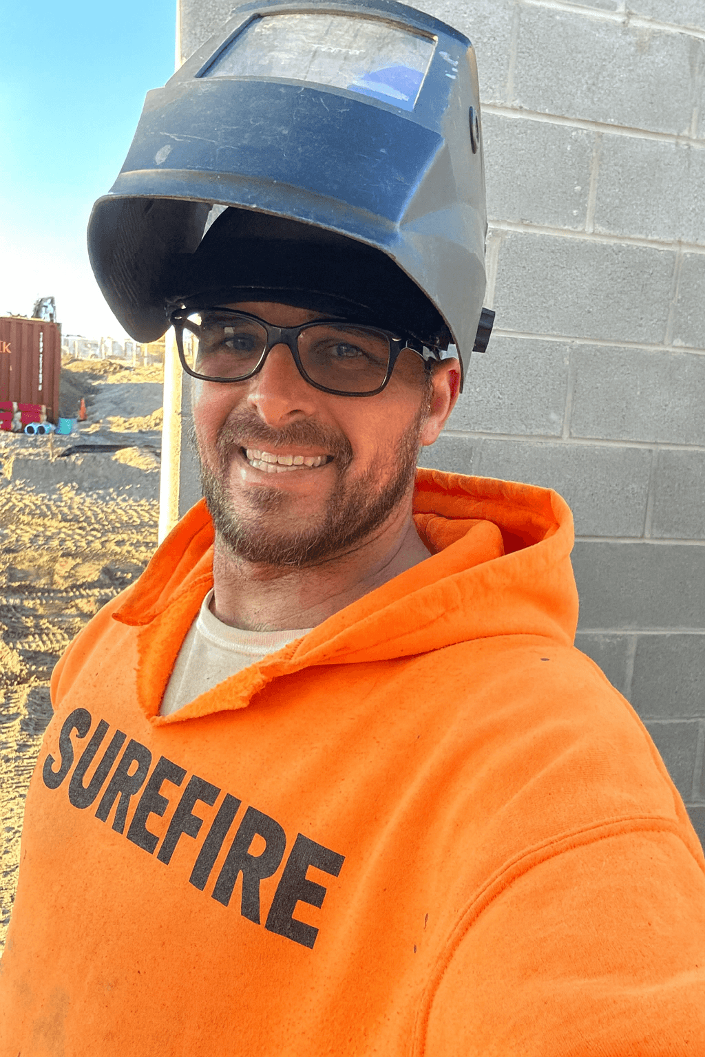 Smiling man in orange Surefire hoodie with a raised welding mask at a construction site.