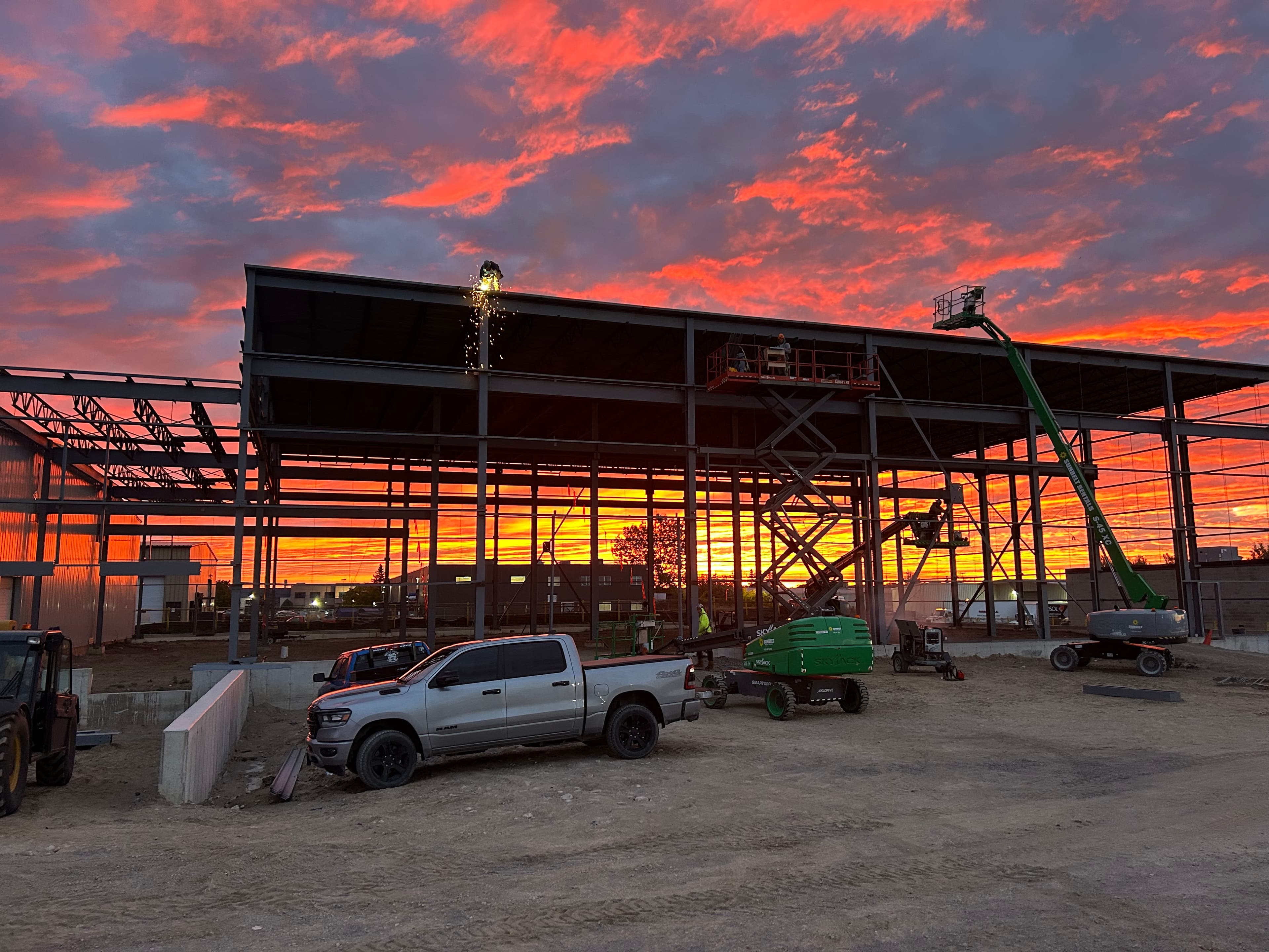 Steel building frame under construction against a fiery orange sunset with sparks and heavy machinery.