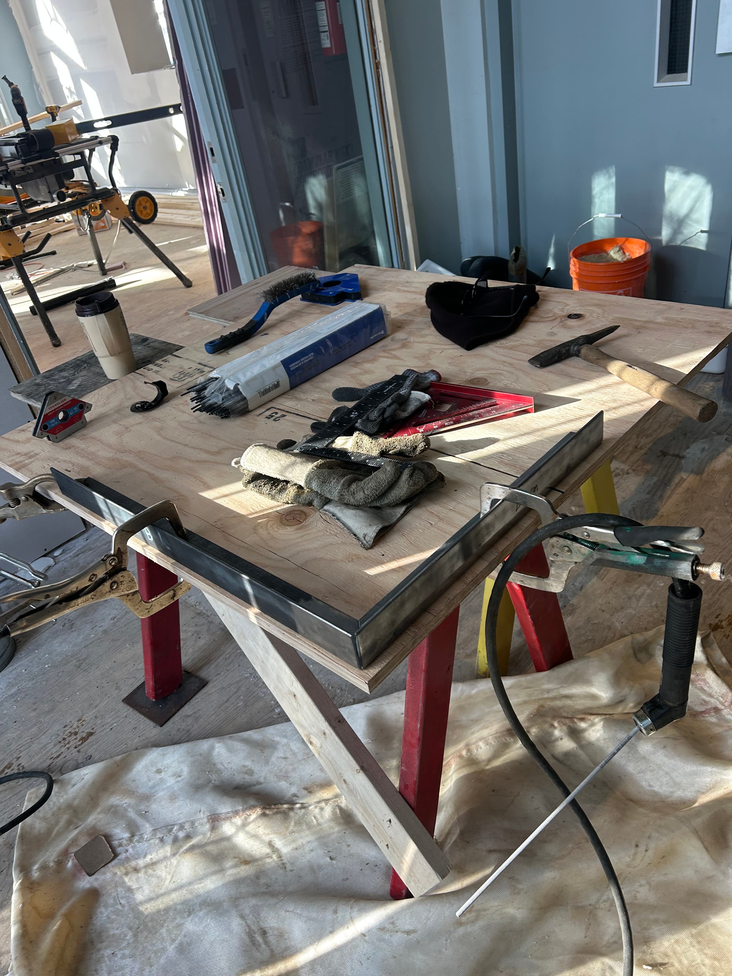 Various welding tools, gloves, and metal pieces scattered across a wooden workbench in sunlight.