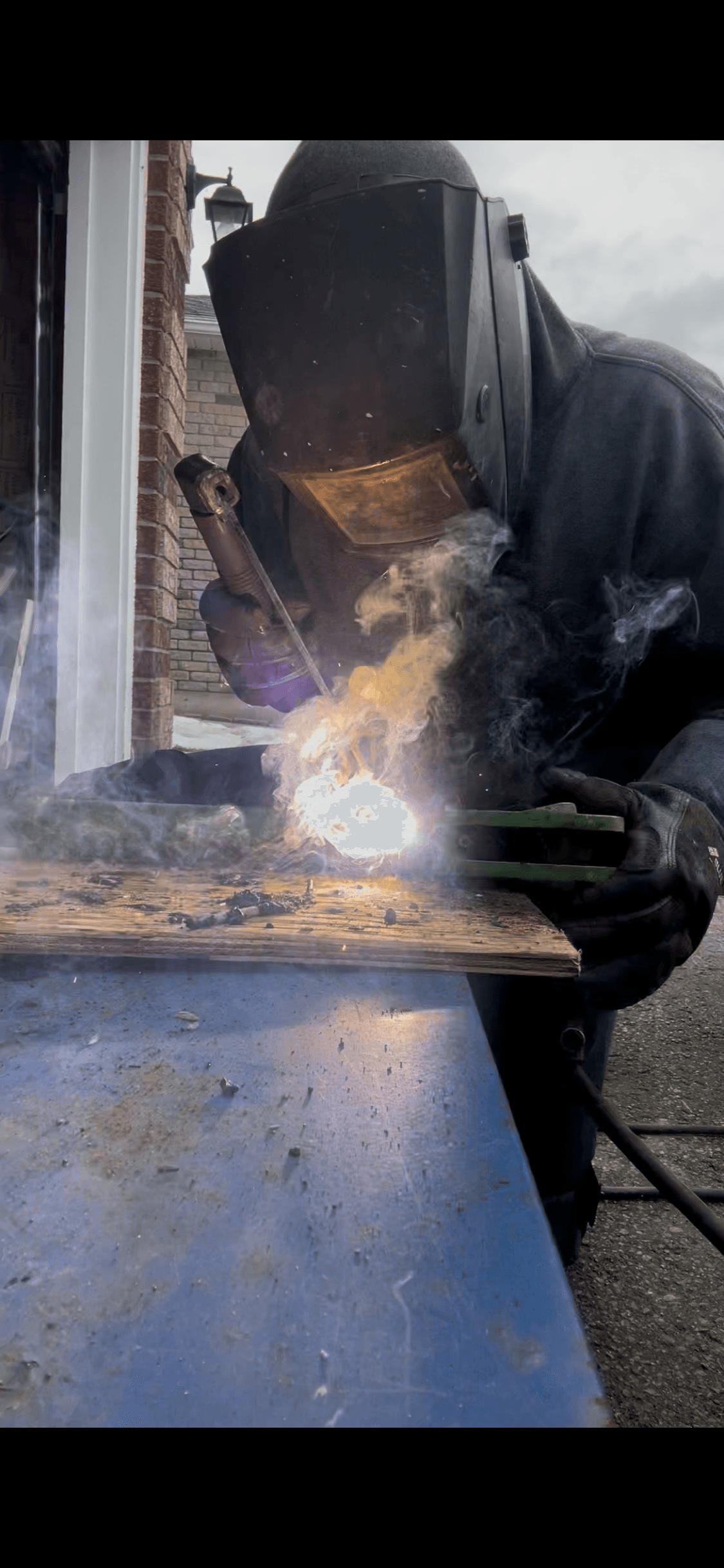 Welder in protective gear works on metal, producing a bright arc and rising smoke.
