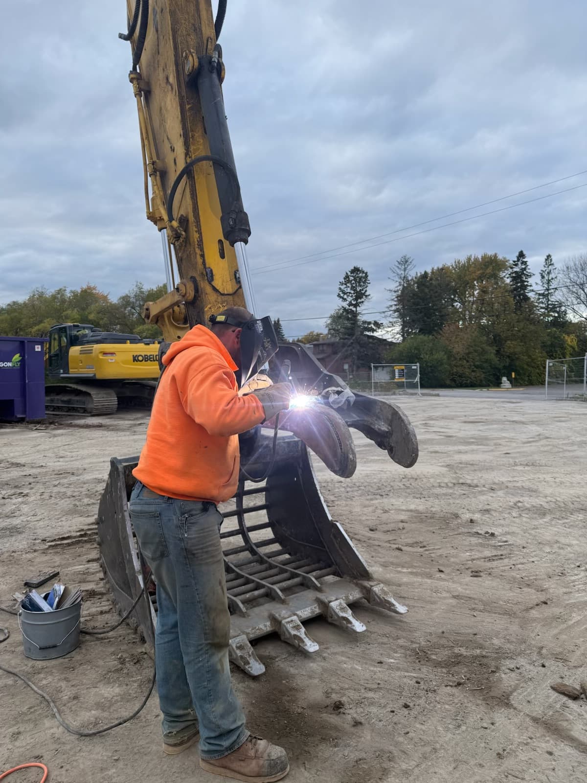 Welder in orange hoodie repairs a yellow excavator bucket at an outdoor construction site.