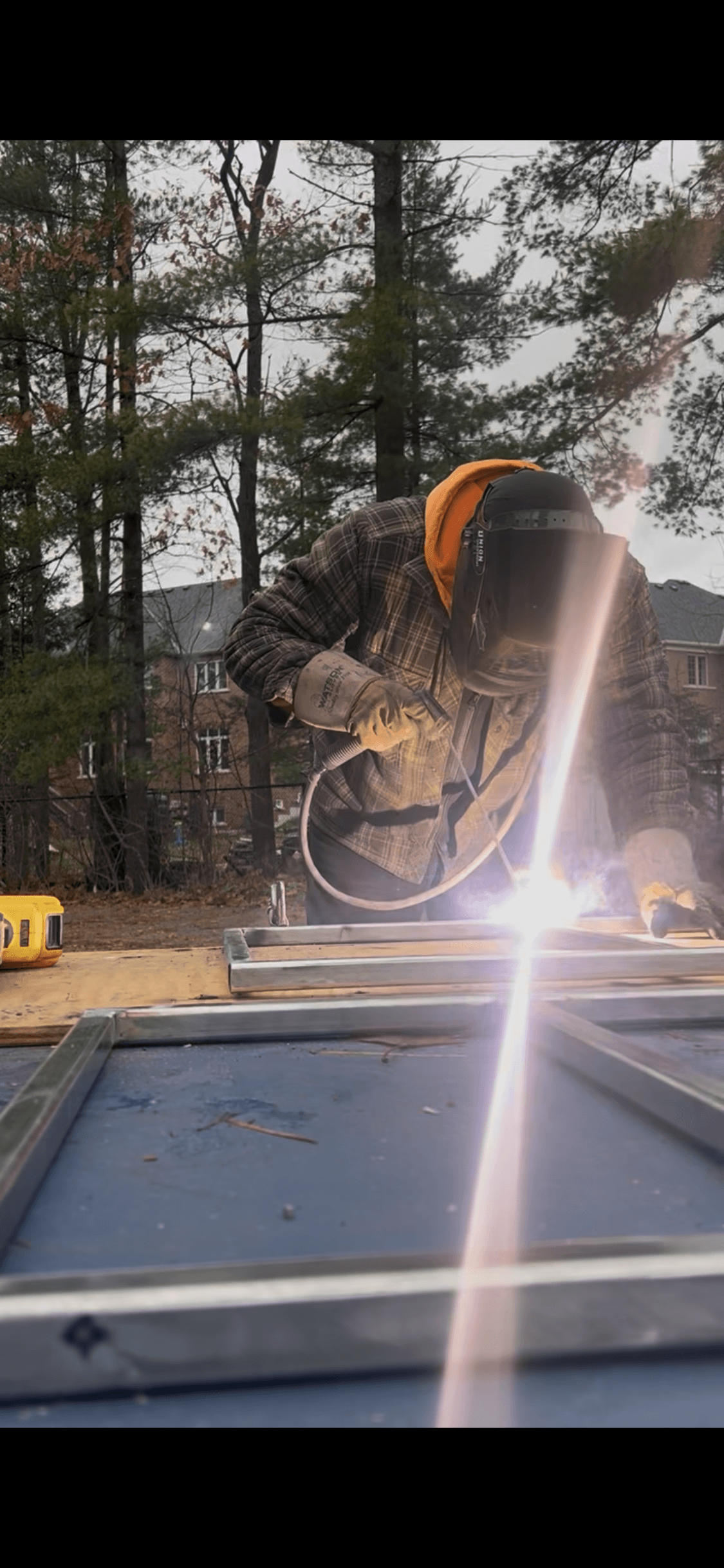 Person in a welding mask welds a metal frame outdoors, creating a bright flash.