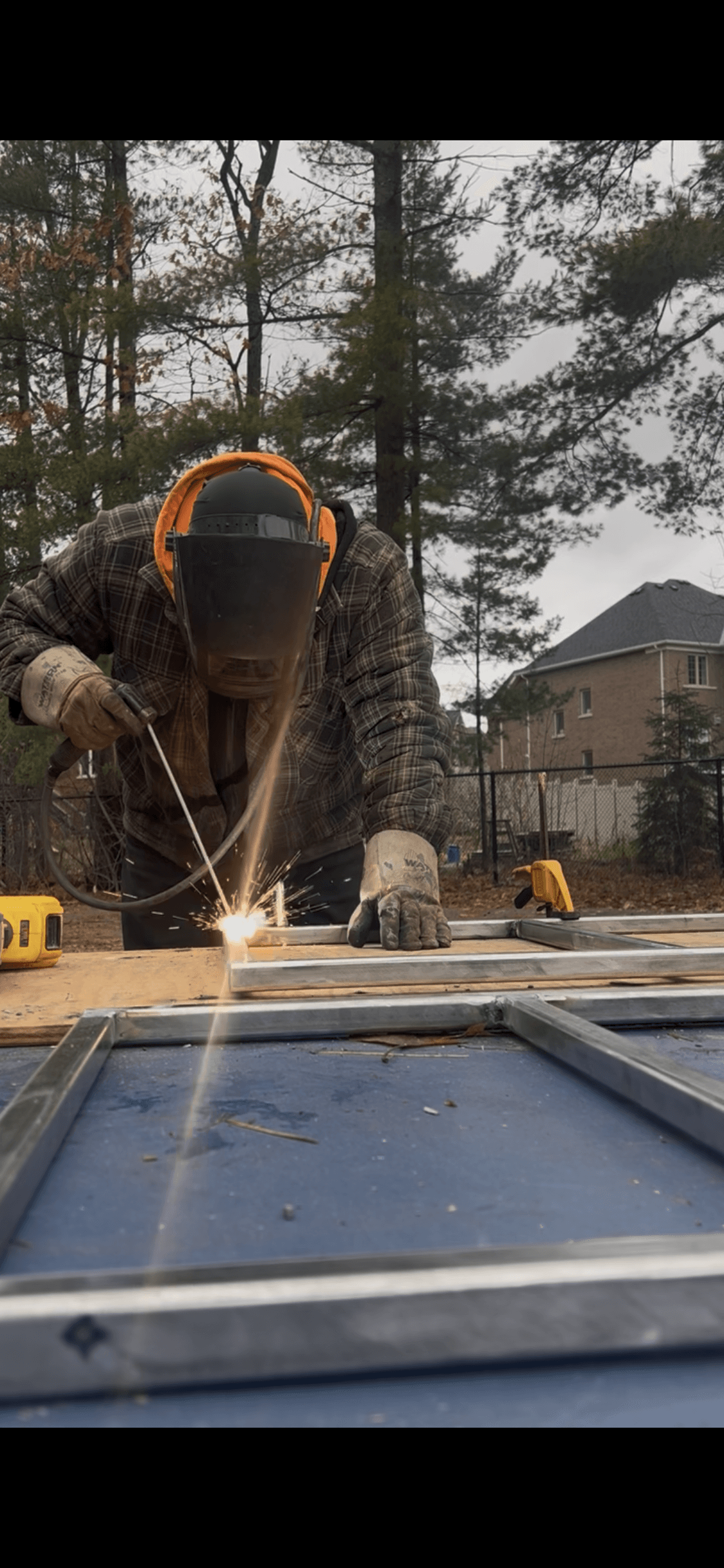 Person in a welding mask and gloves welds metal frames outdoors with bright sparks.